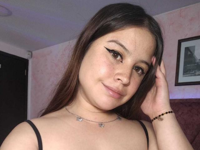 Melani Becker hat langes, braunes Haar, trägt ein schwarzes Top mit Spaghetti-Trägern, hat einen Eyeliner im Winged-Stil, eine Schmetterlingskette und ein Perlenarmband und lächelt sanft in die Kamera..
