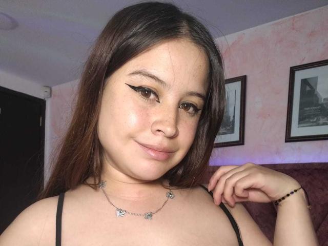 MelaniBecker has brown eyes, black eyeliner, and a slight smile. She's wearing a black top with a star necklace, her hand on her shoulder. Her tits are small.