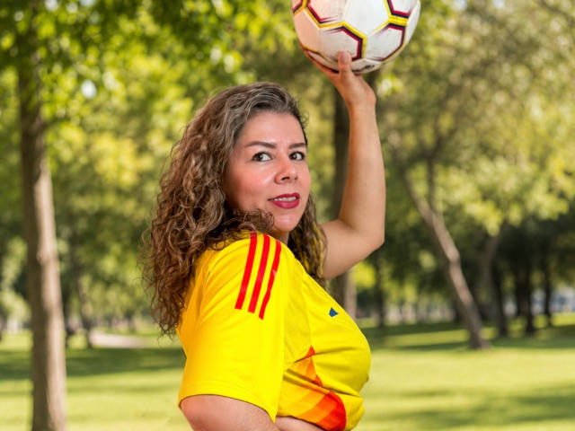 EmiliaMilf com cabelo castanho encaracolado, camisa de futebol amarela com listras vermelhas, segurando uma bola de futebol acima da cabeça, sorrindo, fundo de parque verde, dia ensolarado, corpo atlético, seios médios..