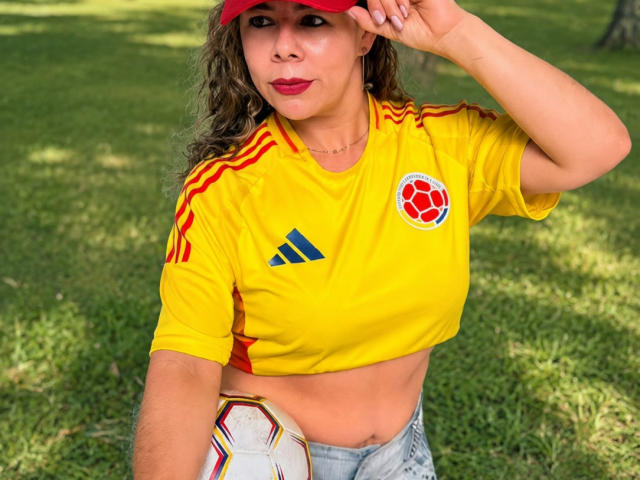 EmiliaMilf con cabello castaño ondulado, gorra roja, camiseta de fútbol amarilla, pelota de fútbol roja, y senos visibles, sosteniendo la gorra, usando jeans, sobre césped verde..