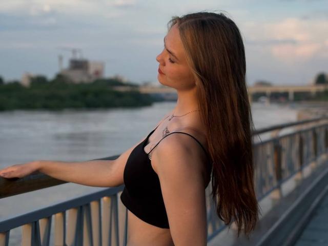 Catherin Bonson mit langen, braunen Haaren, einem schwarzen Sport-BH und einer Halskette, steht auf einer Brücke, blickt zur Seite, hat eine Hand auf dem Geländer, bei Sonnenuntergang..