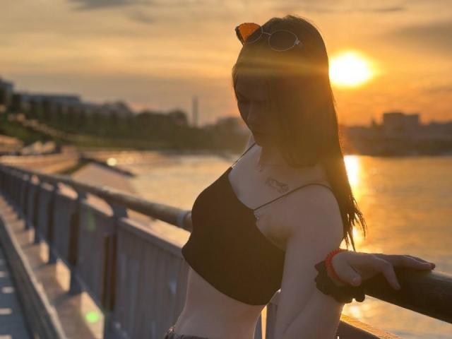Catherin Bonson, avec ses cheveux bruns illuminés par le soleil, un crop top noir, des seins visibles, debout sur un pont, portant des lunettes de soleil sur la tête, un coucher de soleil en arrière-plan, une rivière à droite, un ciel orange..