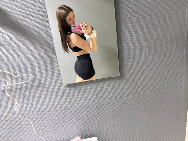 Catherin Bonson, com cabelos castanhos longos, vestindo um top de ginástica preto e shorts pretos de cintura alta, tirando uma selfie no espelho com um celular rosa, mostrando suas nádegas..