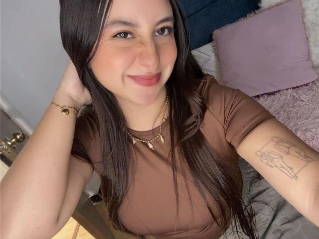 Rosalie Dupont, con lunghi capelli castani, sorridente, indossa una maglia marrone, una collana d'oro e un braccialetto, mostrando un tatuaggio di due donne sul braccio, con la testa appoggiata sulla mano..