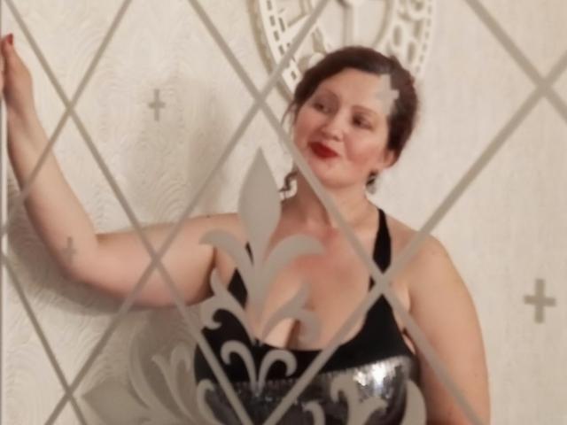 AriaxKiss with red lipstick, black and silver dress, showing tits, smiling, right hand on white patterned wall, brown hair, fair skin, diamond-shaped glass in foreground.