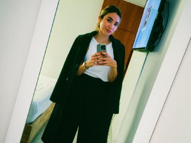 AdharaCooperr stands in front of a mirror, taking a selfie. She's wearing a black blazer, white tee, and black pants, with her brown hair in a ponytail.