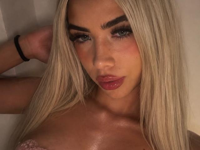 ChristieGarci with blonde hair, blue eyes, and glossy pink lips, looking at the camera, hand behind head, shiny skin, black wristband, and visible cleavage.