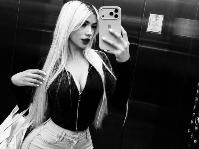 ChristieGarci with long blonde hair, dark lipstick, black zip-up top, showing her big tits, holding phone in elevator, wearing light jeans, reflecting in mirror, looking sexy.
