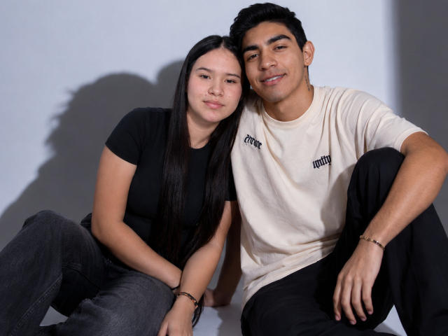 Milo e Mily estão sentados juntos. Milo tem cabelo escuro, pele morena e uma camiseta branca. Mily tem cabelo preto longo, pele clara e uma camiseta preta. Ambos estão sorrindo..