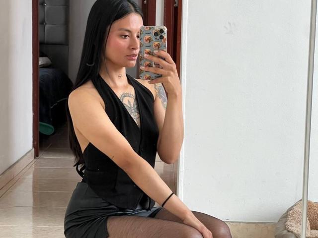NiaWhitee, con cabello largo y negro, vestida con un vestido negro de tirantes y medias transparentes, sentada, sosteniendo un teléfono con una funda estampada con flores, mostrando tatuajes en su pecho..