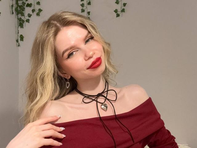 LoveSofiaX with blonde hair, red lipstick, off-shoulder burgundy top, black choker with heart, winking, hand on chest, nails painted white, green vine in background.