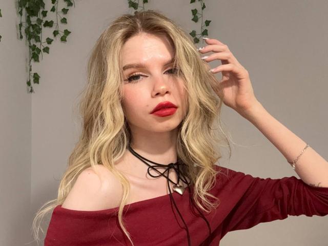 LoveSofiaX with wavy blonde hair, red lipstick, and off-shoulder red top, wearing a black choker, touching her hair, pale skin, and a delicate bracelet on her wrist.