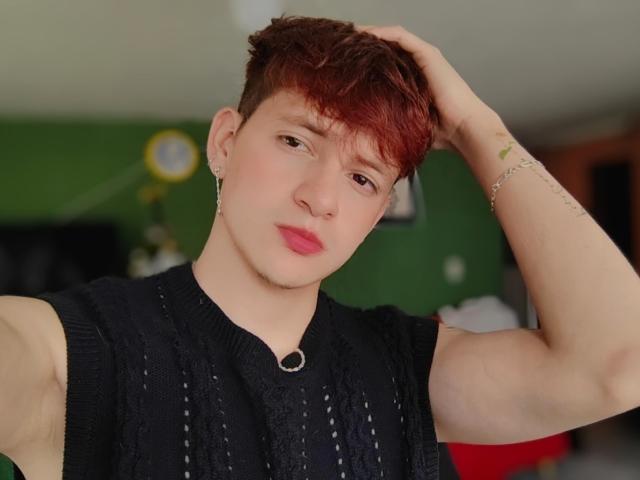 GabyThoms, avec des cheveux teints en rouge, des lèvres roses, un t-shirt noir sans manches, une main dans les cheveux, des boucles d'oreilles, un bracelet, des tatouages, et une expression sérieuse. GabyThoms a une silhouette mince et mignonne..