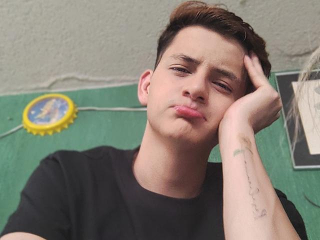 GabyThoms met een fronsende gezicht, een zwart T-shirt, kort bruin haar, de rechterhand op het hoofd, een groen tapijt, een gele ring en een tatoeage op de linkeronderarm..