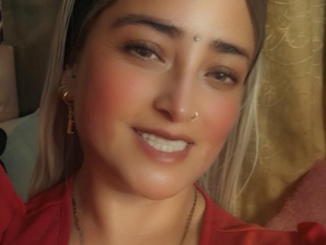 AlessadraJoness with blonde hair, nose ring, and smiling, showing her teeth, wearing a red shirt, and gold earrings, has a small dot on her forehead.