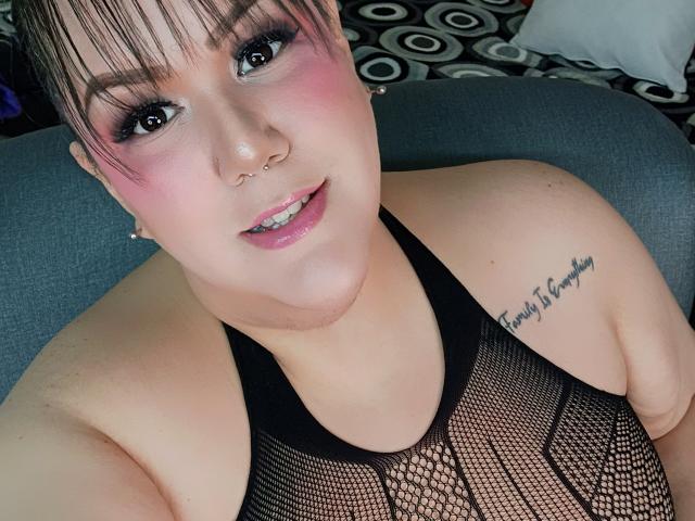 GiiannaMorrys with pink blush, black mesh top, nose ring, and tattoo, showing off her tits and a cute smile, looking at the camera. Short pink hair, white pillow.