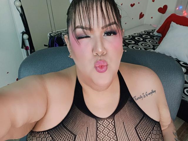 GiiannaMorrys with puffy pink lips, black mesh top, nose ring, and tattoo on right shoulder, winking and sticking out her tongue. She has pink eyeshadow and dark hair.