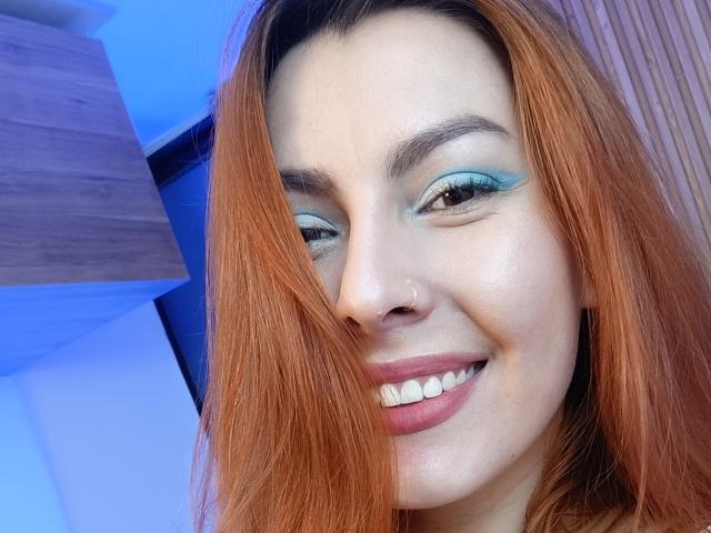 AnnyCrafforts Gesicht mit roten Haaren, blauem Lidschatten, einem Lächeln, weißen Zähnen und einem niedlichen Nasenring, in einem Selfie. Ihre Brüste sind nicht sichtbar..