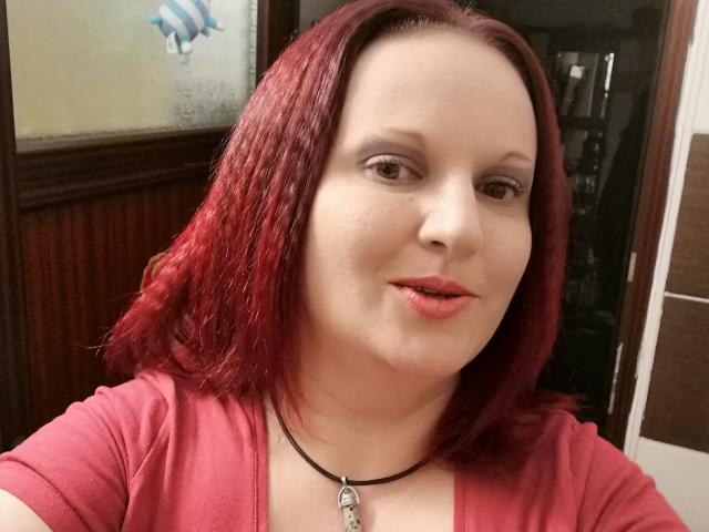 LadyAlma a les cheveux roux, une peau claire, un petit nez, un maquillage discret, des lèvres roses, et elle porte un haut rouge avec un collier noir. Ses seins sont de taille moyenne, et elle regarde l'appareil photo..