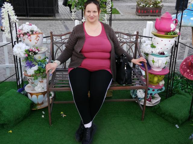 LadyAlma, avec de grandes seins, un haut rose, un pantalon noir, un cardigan marron, un sac noir, assise sur un banc, entourée de grandes tasses à thé, de fleurs blanches, d'herbe verte, souriante, avec des cheveux foncés, devant une clôture blanche..