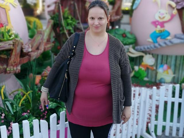 LadyAlma mit einem runden Bauch, die ein pinkes Oberteil, schwarze Hosen und einen grauen Pullover trägt, steht mit einer schwarzen Tasche in der Hand, blickt in die Kamera und lächelt..