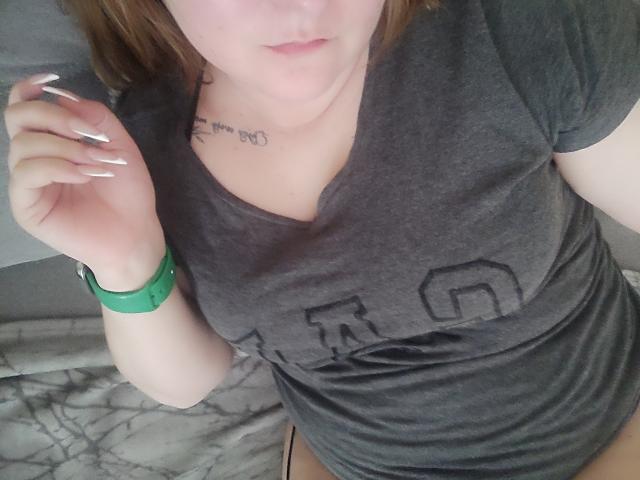 MinistraX in a gray tee, green wristband, and long white nails, with a tattoo under her chin, looking down. Her boobs are medium-sized and she has red hair.