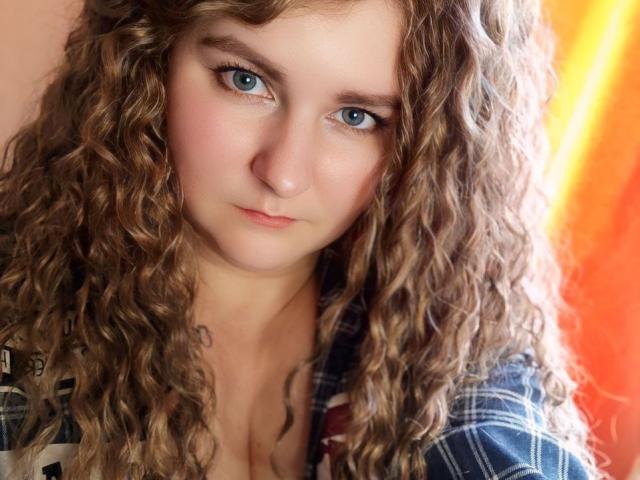 MinistraX with curly brown hair, blue eyes, and a slight pout, wearing a plaid shirt, showing cleavage, looking directly at the camera with a neutral expression.