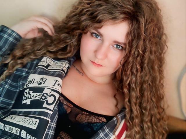 MinistraX has curly brown hair, blue eyes, and a slight smile. She's wearing a plaid shirt over a black lace top, exposing her tits. Her skin is fair.