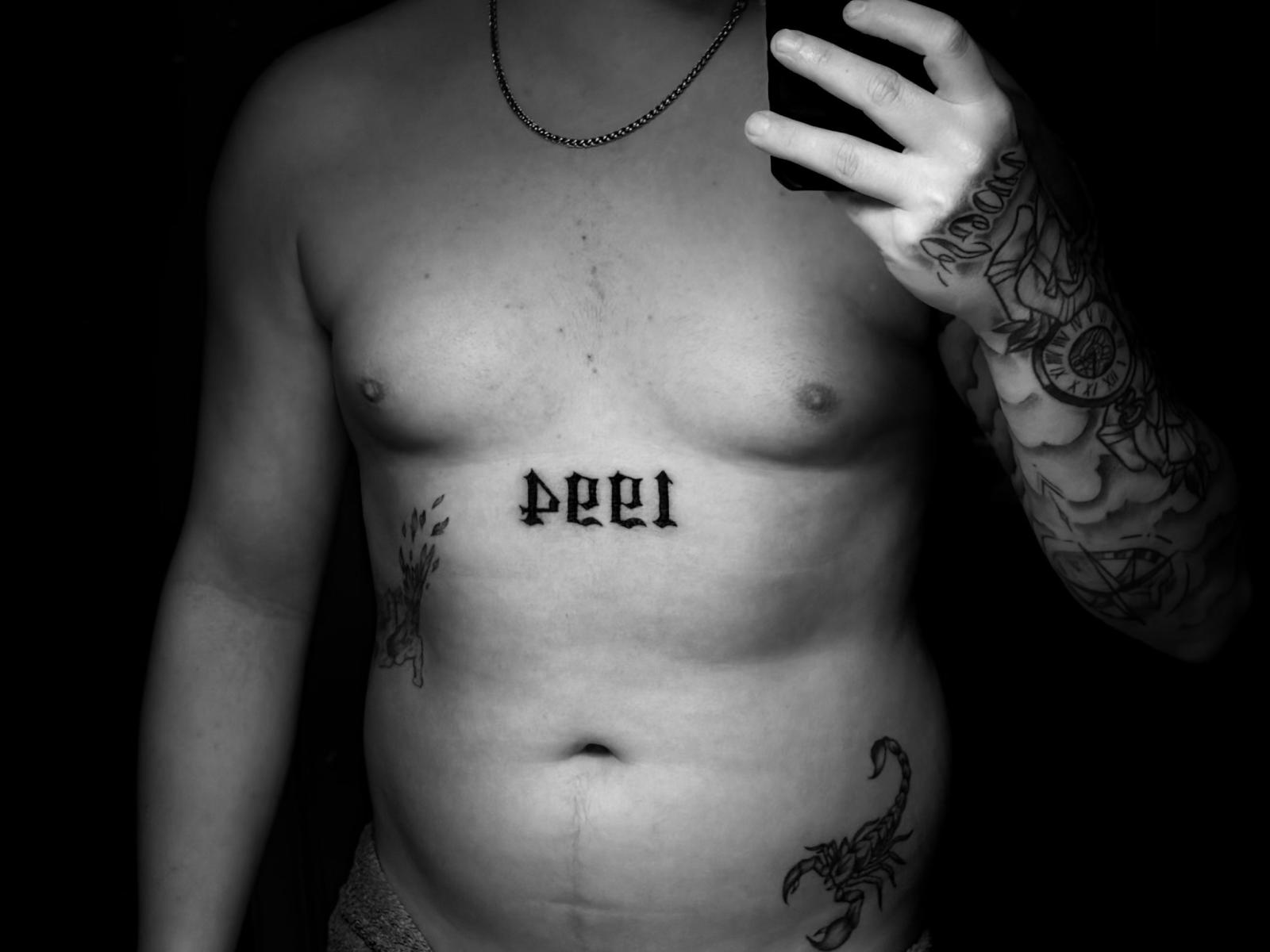 Il petto nudo, leggermente paffuto, di Xonix69, con un tatuaggio con la scritta "001" in lettere gotiche nere, che tiene un telefono, un braccio tatuato e un tatuaggio sulla parte inferiore dell'addome, su uno sfondo scuro..