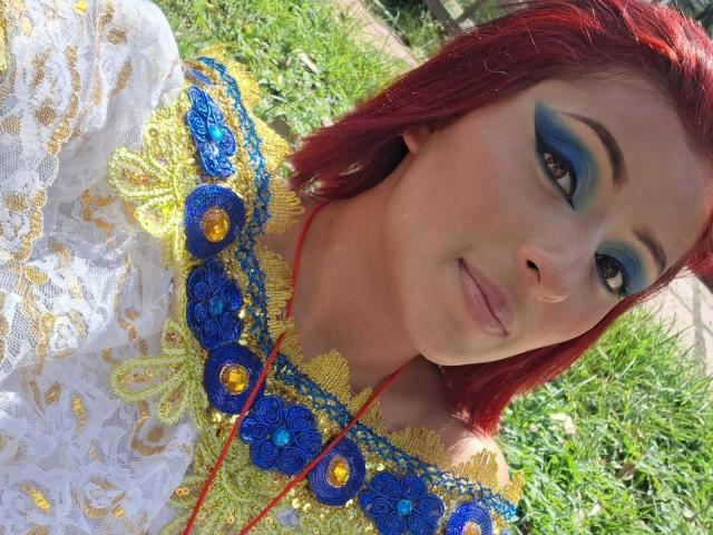 LizBonnet, avec des cheveux roux, un maquillage bleu et jaune pour les yeux, une robe blanche en dentelle et un collier floral coloré, assise sur de l'herbe verte, souriant à la caméra. Les seins ne sont pas visibles..