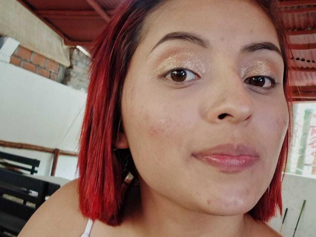 Le visage de LizBonnet, avec sa coupe courte teintée en rouge, ses yeux marron brillants et ses lèvres roses et brillantes, présente de légères imperfections sur sa joue gauche. Elle porte un haut blanc, et un toit rouge est visible en arrière-plan..