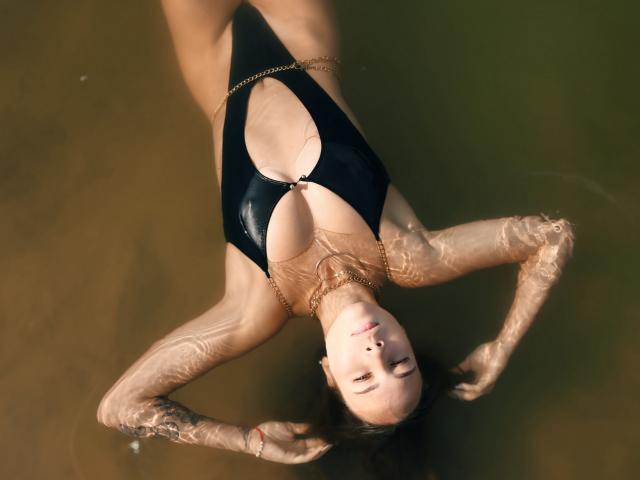 AmmaLoyXX floating in water, wearing a black, shiny one-piece with a deep cleavage, arms above head, gold chain necklace, wet skin, dark hair, relaxed expression.