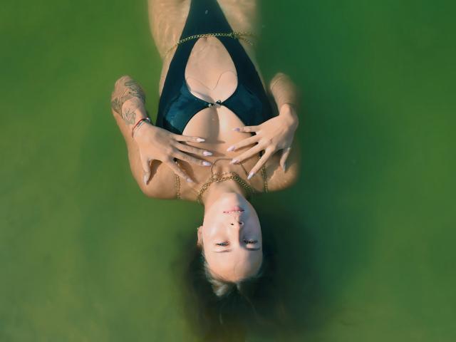 AmmaLoyXX with tattoos, lying in green water, wearing a black one-piece with a deep cleave, hands on chest, gold chain, red bracelet, and white nail polish.