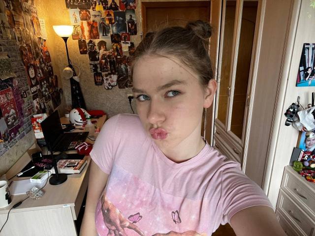 ShiraWolny with pouty lips, wearing a pink shirt with pink butterfly prints, brown hair in two buns, in a cluttered room with photos on the wall.