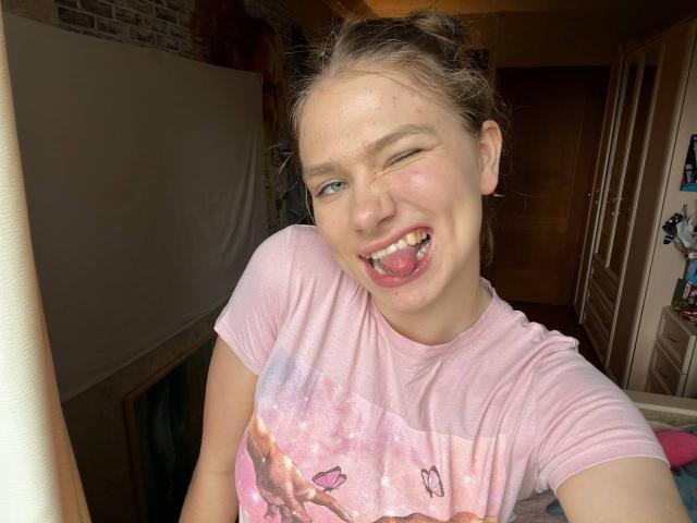 ShiraWolny, with a goofy smile and tongue out, winks at the camera, wearing a pink tie-dye shirt, has light skin, and brown hair in a messy bun.