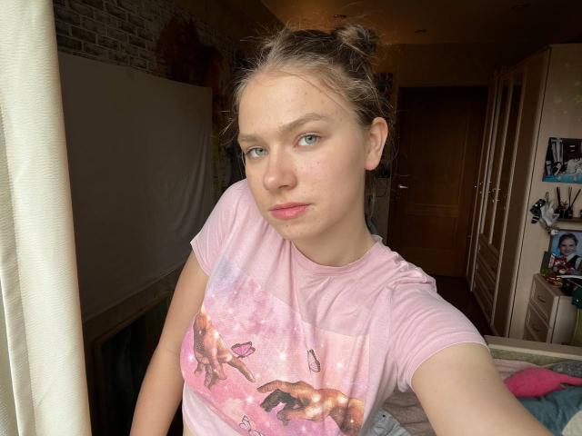 ShiraWolny has light skin, blue eyes, and pink lipstick. She's wearing a pink tee with a unicorn print, her hair is in two buns, sitting by a window.