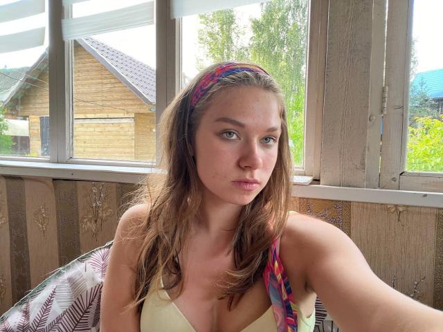 ShiraWolny with medium tits, wearing a yellow bra and colorful headband, has long brown hair, green eyes, and a serious expression, sitting in a wooden room with a window.