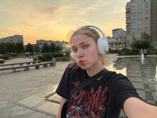 ShiraWolny with puffy lips, wearing white headphones, black graphic tee, and a strap across her chest, standing in a city square at sunset, her ass slightly visible.