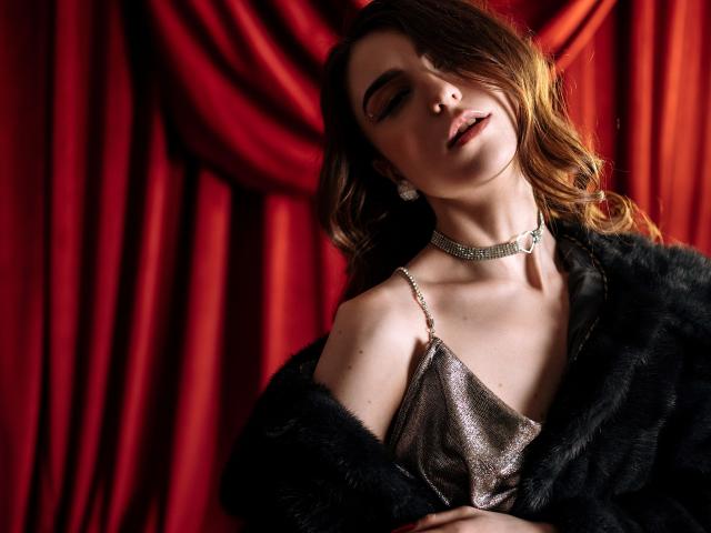 MelanieFoster with eyes closed, wearing a sparkly silver dress, black fur coat, and diamond choker, her head tilted back, looking sexy and relaxed. Red curtain in background.