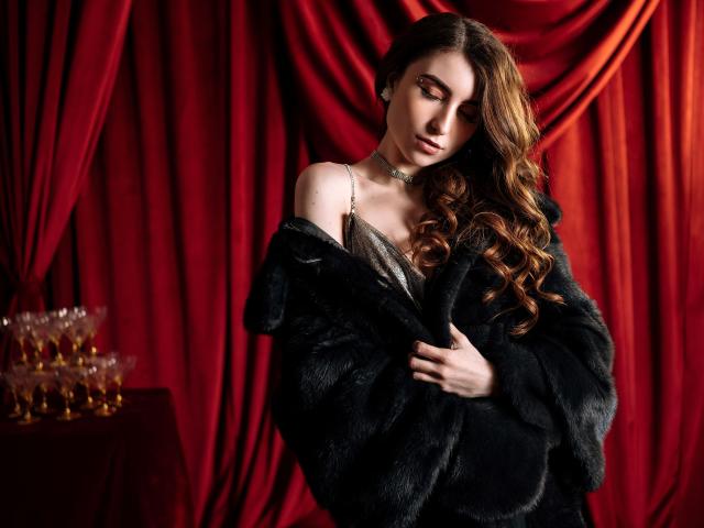 Melanie Foster, com longos cabelos castanhos ondulados, vestindo um casaco de pele preto, que revela parcialmente lingerie prateada e um colar, olhando para baixo com uma expressão sedutora e séria..