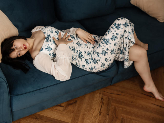 ChrissieCandy liegt auf einem blauen Sofa, trägt ein Blumenkleid, ihre Brüste sind entblößt, ihre Hand liegt auf ihrem Genital, und ihre Beine sind gekreuzt, auf einem Holzboden mit Fischgrätmuster..