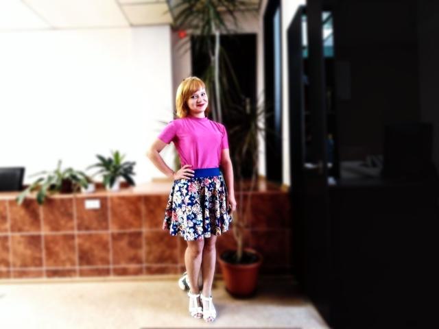 KaterinCold, com cabelo ruivo, blusa rosa, saia azul com estampas florais, saltos brancos, em pé com a mão na cintura, sorrindo, em um quarto com piso de azulejos e plantas em vasos..
