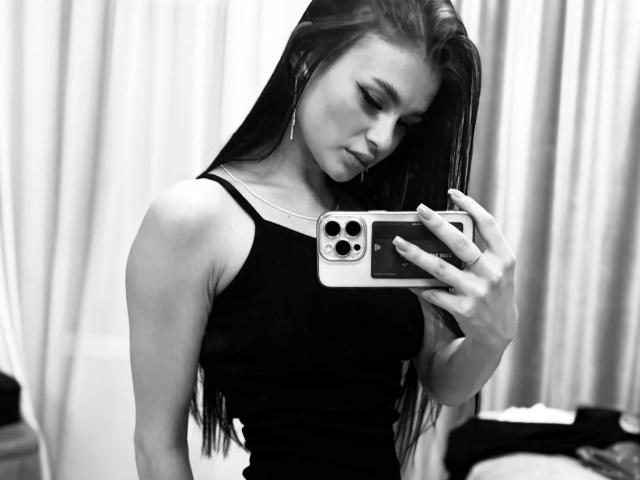 Photo en noir et blanc de Mickie Silkwood, avec de longs cheveux foncés, portant un débardeur noir, prenant un selfie devant un miroir avec un téléphone, montrant ses seins et un léger sourire..