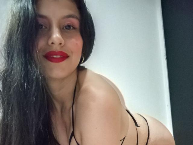 MadissonFontaine with long black hair, red lipstick, smiling, wearing a black strappy lingerie top, showing her bare back and a hint of her ass.