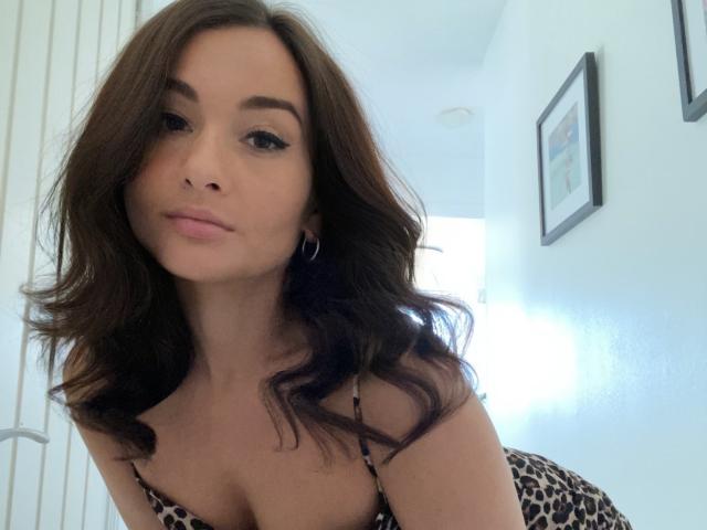 Dionisia with wavy brown hair, pink lips, hoop earrings, and leopard-print lingerie, leaning forward, showing her tits, in a bright, white room.