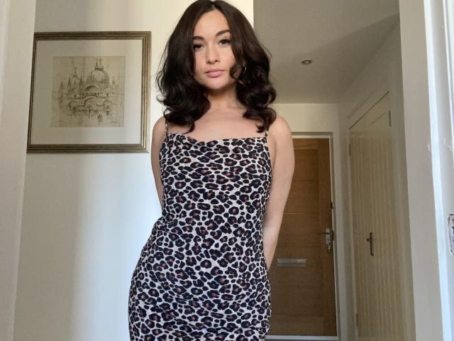 Dionisia has wavy black hair, wearing a leopard-print dress, standing with hands behind her back, showing her ass and small tits, in a white-walled hallway.