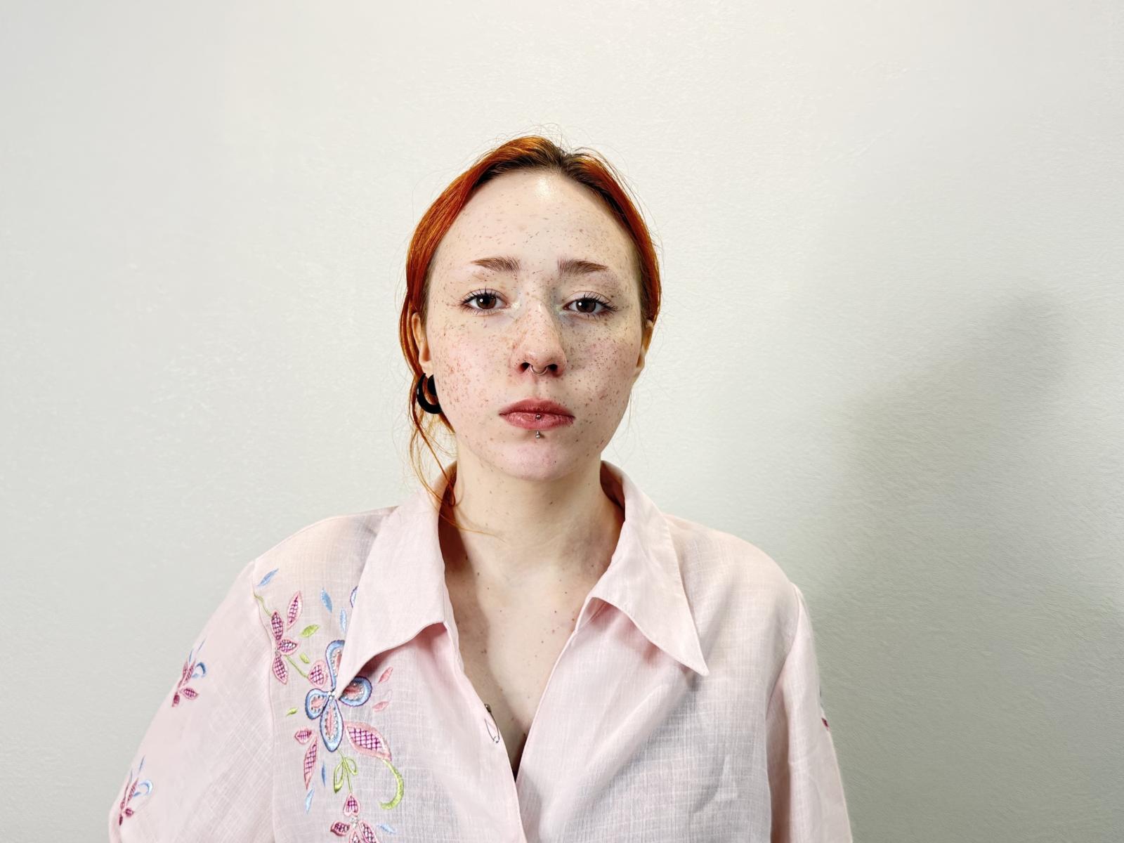 BariBenafield has red hair, freckles, and a pink button-up shirt with colorful floral embroidery. She's looking directly at the camera with a neutral expression.