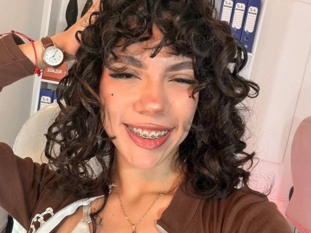 Angelica Hill, con i capelli castano ricci, sorride mostrando denti con una griglia metallica, indossa una felpa marrone con zip, un braccialetto rosso, una collana dorata e ha una mano tra i capelli, rivelando un piccolo neo sulla guancia destra..