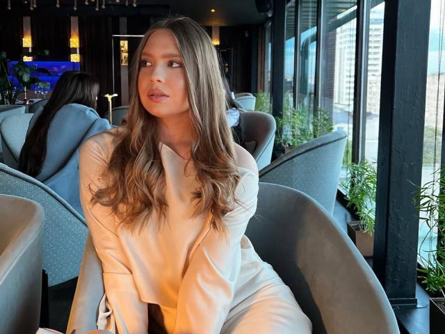 Margrett Munley, con lunghi capelli castani ondulati, indossa un abito bianco a maniche lunghe, è seduta su una sedia grigia, guarda di lato, in un moderno caffè pieno di luce solare..
