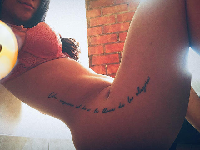 Os seios de LaraMancini estão em um sutiã de renda rosa, mostrando uma tatuagem no lado direito, logo acima da vulva. O bumbum dela está ligeiramente levantado e ela está sorrindo..